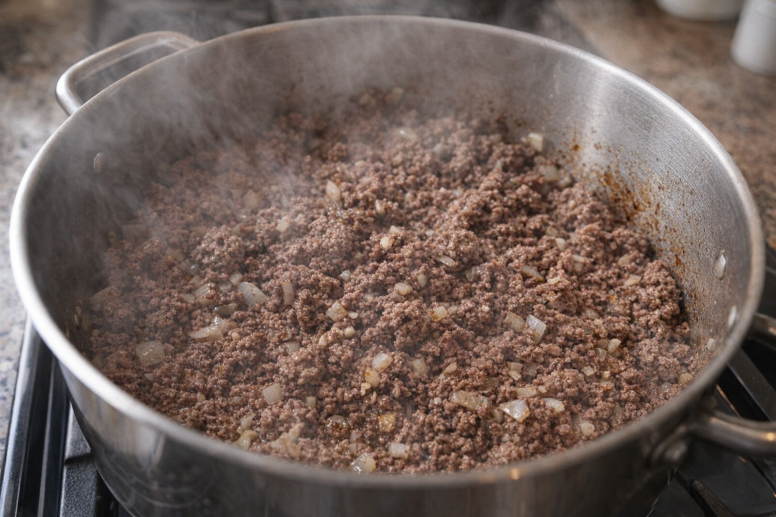 An overhead shot of ground beef being browned in a large pot on a stovetop, with visible steam and browning on the meat. Onions and garlic are visible in the pot as well